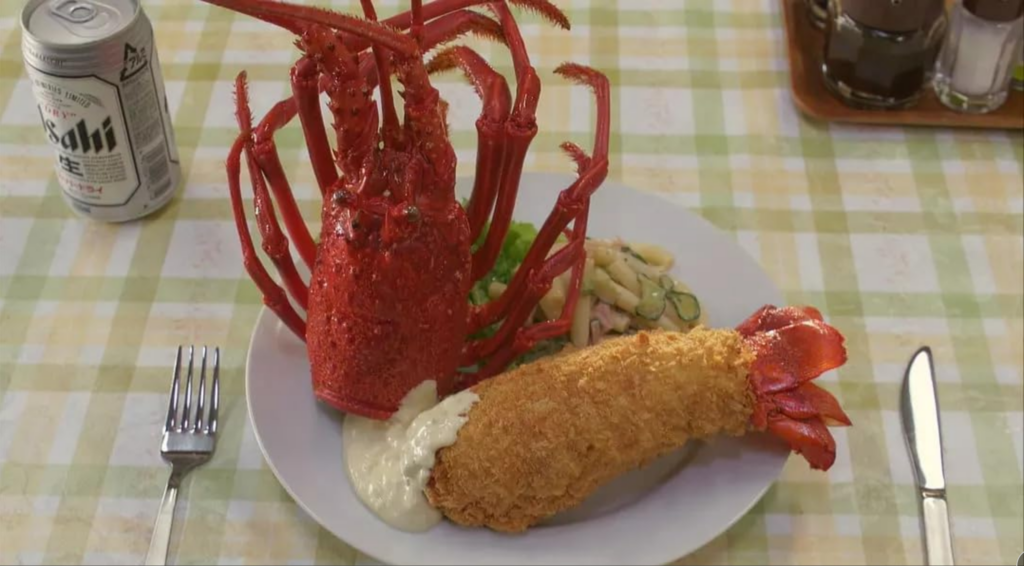 大きな伊勢海老のフライの料理がプレートに盛り付けられ、その横にビール缶が置かれている食事のシーン